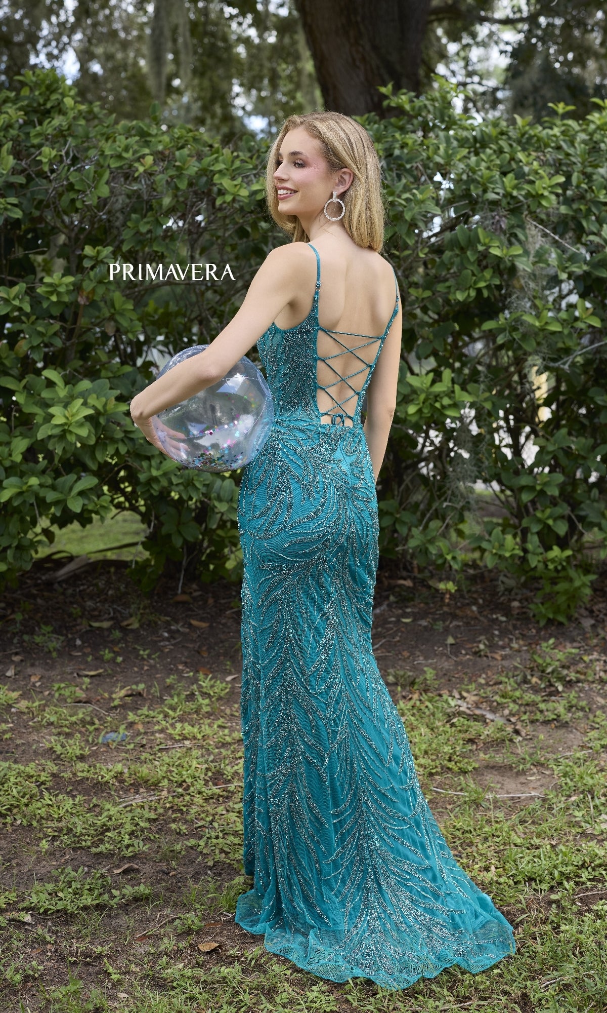 A woman stands on grass outdoors holding a clear confetti balloon, wearing the Primavera 4310 Scoop-Neck Long Beaded Prom Dress. Lush green bushes form the background.